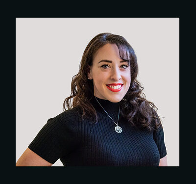 photo of Jocelyn Rosnick, woman with long curly brown hair wearing a black short sleeved top and a round pendant necklace