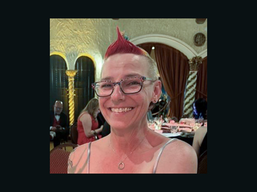 Photo of KD Donaldson, person with a rainbow mohawk and glasses, wearing a dress.