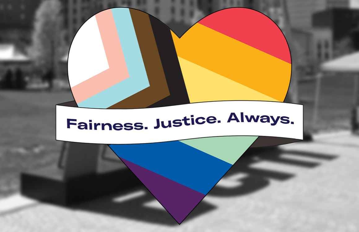 Pride flag in the shape of a heart with a banner across the front that says "Fairness. Justice. Always." on a blurred, black and white background photo.
